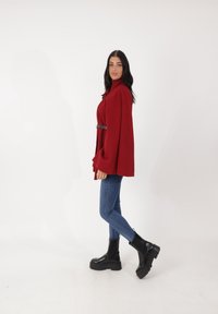 Mantello rosso con collo alto e maniche arrotolate, stretto in vita con una cintura nera. Indossato sopra jeans blu e stivali neri con plateau alla caviglia.