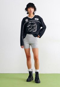 Juodas sportinis marškinėliai su baltais akcentais ir numeriu 23, derinami su pilkais šortais ir juodais raišteliais batais. Lygus fonas ir žalias grindis.