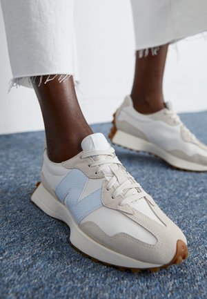 Osoba w białych i beżowych trampkach z jasnoniebieskim logo "N" oraz białych spodniach z postrzępionym dołem stoi na niebieskim dywanie.