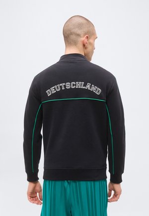 Homme à la tête rasée portant une veste noire avec le texte "DEUTSCHLAND" au dos et un pantalon rayé vert, tourné dos à la caméra.
