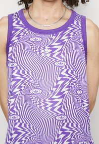 Lila träningslinne med en vågig randig mönstring i vitt och marinblått, med en liten Adidas-logotyp i mitten. Slät tyg och ärmlös design.