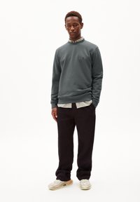 Sweatshirt gris avec des poignets côtelés, superposé sur une chemise à carreaux clairs. Un pantalon noir et des baskets beiges avec des accents pastel complètent le look.