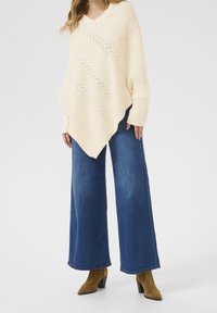 Beige stickad poncho med V-ringning, prydd med ett sicksackmönster. Kombinerad med vida blåjeans och bruna klackboots.