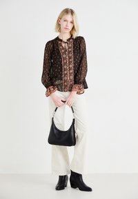 Blusa nera trasparente con motivi floreali e polsini arricciati, abbinata a pantaloni larghi color crema e stivaletti neri con tacco, mentre tiene una borsa nera.