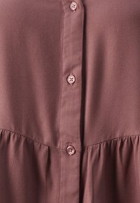 Camicia button-up color rosa bruno con una superficie testurizzata, dotata di cinque bottoni sul davanti e una cucitura arricciata nella parte inferiore.