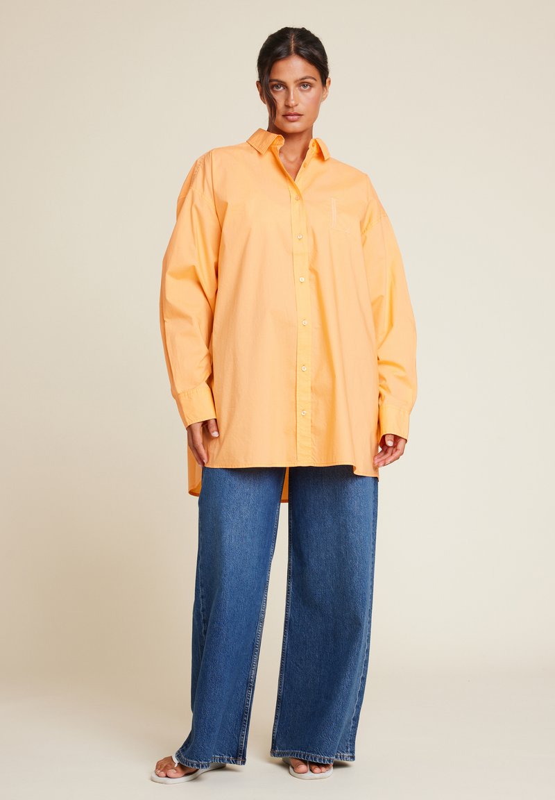 Overdimensjonert oransje skjorte laget av bomull, med lange ermer, krage og en brystlomme, kombinert med vide blå denimjeans.