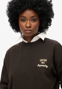 Felpa marrone con colletto a girocollo, con testo ricamato bianco "UNION MFG Superdry". Materiale in cotone con una texture morbida.