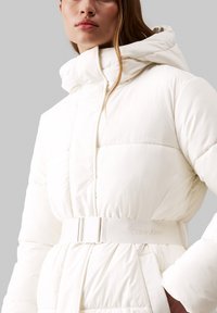 Balta pūkinė striukė su kapišonu, su diržu per liemenį ir subtiliu Calvin Klein ženklu. Lygi tekstūra ir siuvinėtas dizainas.