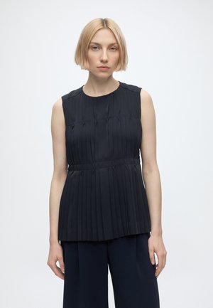 Giovane donna con capelli biondi corti che indossa un top nero senza maniche plissettato e pantaloni neri, in piedi davanti a uno sfondo bianco semplice.
