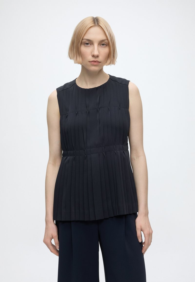 Jeune femme aux cheveux courts et blonds portant un haut noir plissé sans manches et un pantalon noir, debout devant un fond blanc uni.
