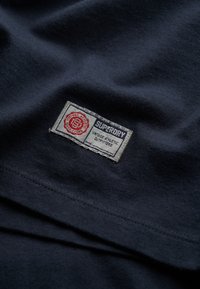 Camiseta de algodón color navy con un parche rectangular que lleva texto en rojo y azul, cosido a lo largo del borde inferior. Textura suave con un ligero brillo.