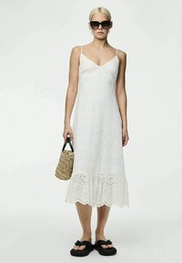 Mulher usando um vestido sem mangas branco com rendas, sandálias pretas, óculos de sol grandes e segurando uma bolsa de palha trançada.