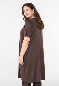 Smaidoša sieviete ar viļņainiem brūniem matiem valkā vaļīgu, ceļgalu garuma brūnu kleitu ar īsām piedurknēm un melnas zeķubikses, stāvot pret vienkāršu fonu.