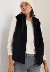 Äkta päls ärmlös väst i marinblått med huva och dragsko, tillsammans med en krämfärgad långärmad skjorta och grå jeans. Slät textur.