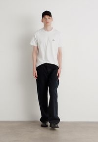 Hvid bomulds t-shirt med et lille grafisk tryk, sorte afslappede bukser og sorte sko. Modellen bærer en sort baseballcap.