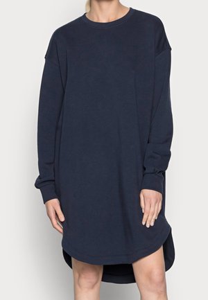 Kvinde iført en langærmet, navyblå oversized sweatshirtkjole med rund halsudskæring og let buet kant.