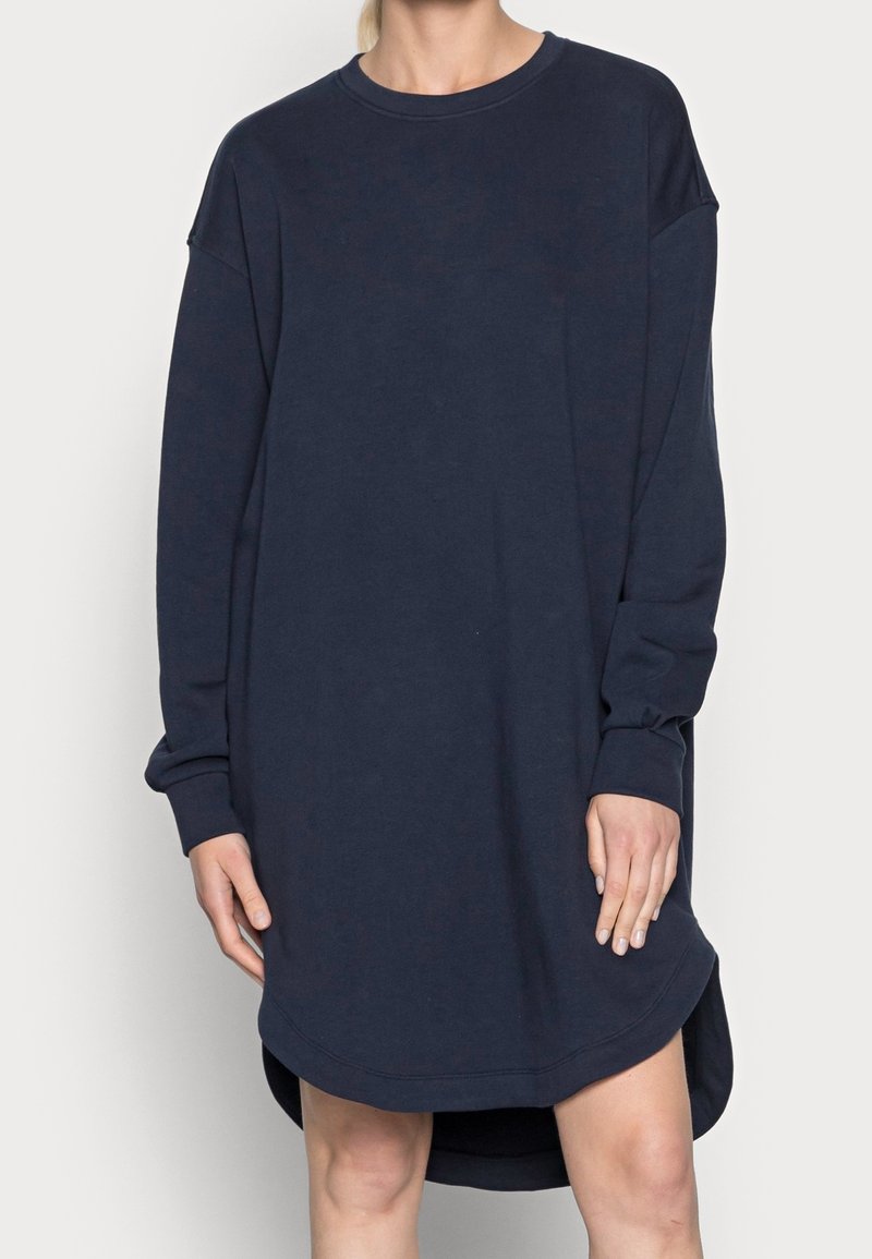 Femme portant une robe sweat-shirt oversize bleu marine à manches longues avec un col rond et un ourlet légèrement courbé.