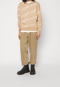 Béžová mikina s bílým grafickým textem, doplněná volnými manšestrovými kalhotami. Černé šněrovací boty završují outfit.