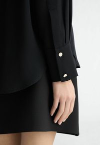 Black blouse with a smooth texture and buttoned cuffs, paired with a black skirt. The design features a relaxed fit and a subtle sheen.