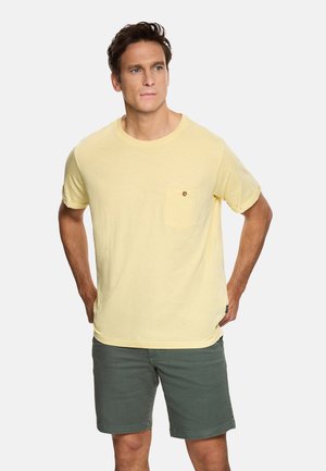 Hombre vestido con una camisa de manga corta color amarillo claro con un bolsillo en el pecho y pantalones cortos de color verde oliva, de pie con las manos detrás de la espalda.