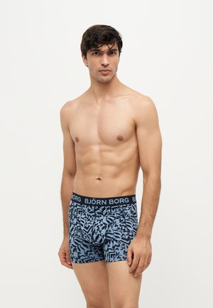 Skjorteløs mand med kort mørkt hår iført blå og sorte mønstrede boxershorts med "BJÖRN BORG" taljebånd står mod en ensfarvet baggrund.