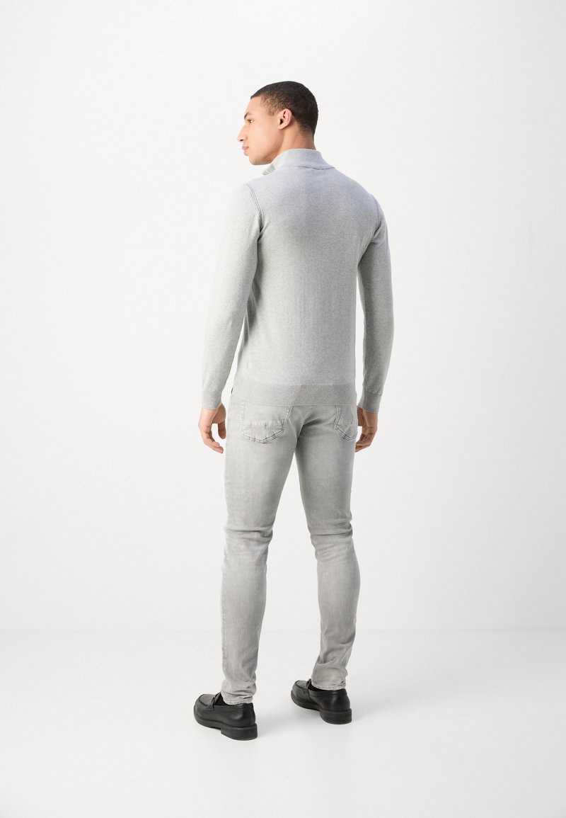 Maglione grigio a maglia con colletto ripiegato, abbinato a jeans skinny grigi chiari e scarpe nere, visto da dietro.