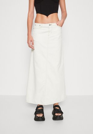 Femme portant une jupe maxi blanche taille haute, un crop top noir et des sandales à plateforme noires avec des brides, debout devant un fond blanc.