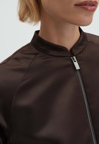 Bruine satijnen bomberjas met een opstaande kraag, voorzien van een volledige ritssluiting en een gladde textuur. Close-up van het bovenste deel.