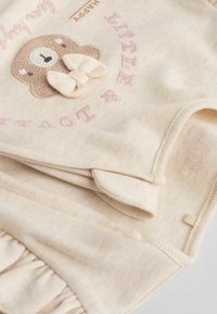 Conjunto de tela suave beige que presenta un oso bordado con un lazo, texto que dice "Pequeño y Querido" y un dobladillo con volantes.