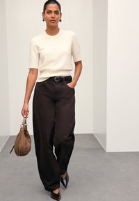 Haut court à manches courtes en maille crème clair, pantalon large marron foncé, ceinture noire, chaussures noires à bout pointu avec un sac à main en daim marron.