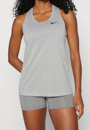 Femme portant un haut de sport gris sans manches Nike et un short de sport gris assorti, debout avec une main derrière le dos.