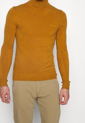 Homme portant un pull à col roulé jaune moutarde et un pantalon beige, debout devant un fond blanc, les mains détendues le long du corps.