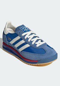 Modrá sportovní teniska s bílým designem tří pruhů, texturovaným jazykem a gumovou podešví. Vyrobena z kůže a semiše s vyztuženou špičkou.