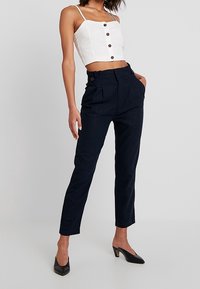 Bílý krátký top s knoflíky a tenkými ramínky, zkombinovaný s kalhotami s vysokým pasem v námořnické modři a černými vysokými podpatky.