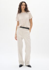 Beige overhemd met korte mouwen, gecombineerd met lichte linnen broeken en een zwarte riem. De outfit heeft een ontspannen pasvorm en soepele texturen. Zwart puntige schoenen.