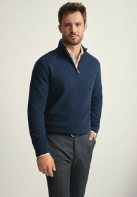 Granatowy sweter rozpinany z teksturowanym wzorem dzianiny, z kołnierzykiem na ćwierć, ściągaczami na rękawach, zestawiony z szarymi spodniami i białą koszulą pod spodem.