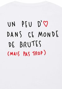 Balta kokvilnas T-krekls ar melnu un sarkanu tekstu uz muguras. Teksts: "UN PEU D'❤️ DANS CE MONDE DE BRUTES (MAIS PAS TROP)" dažādos fontos.