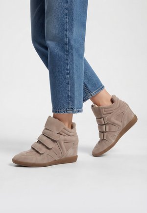 Persona que lleva zapatillas beige con cuña, tres tiras de velcro y vaqueros azules sobre un fondo liso.