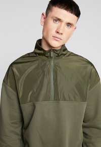 Giacca pullover verde oliva con colletto alto, chiusura a mezzo zip e trame di tessuto opache e lucide a contrasto.
