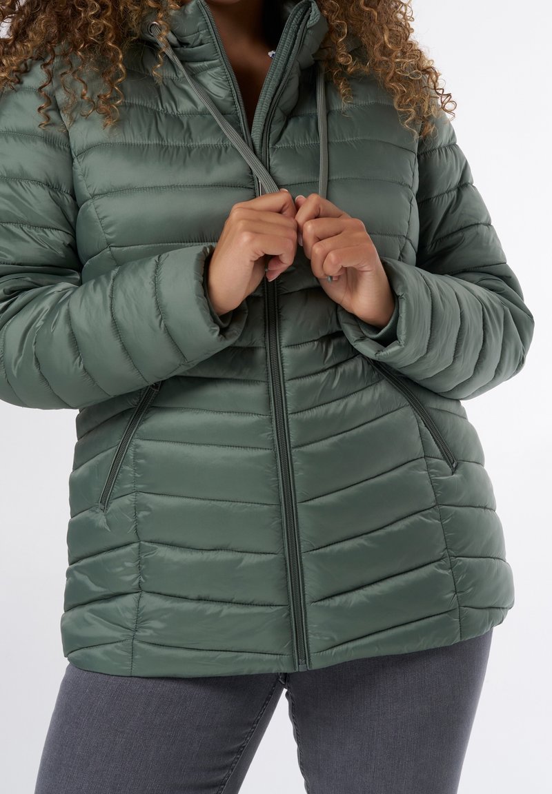 Personne aux cheveux bouclés ajustant les cordons de serrage d'une veste matelassée verte, portant un pantalon gris foncé, sur un fond blanc uni.