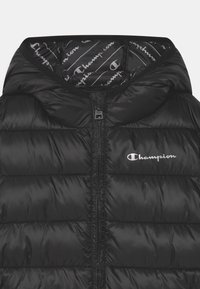 Czarna kamizelka puchowa z kapturem, posiadająca poziome pikowane sekcje, pełny zamek błyskawiczny oraz biały logo "Champion" na lewej części klatki piersiowej.