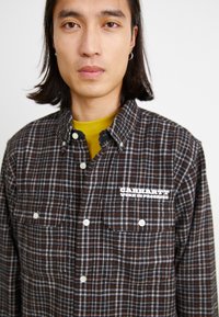 Chemise en flanelle noire, orange et blanche à carreaux ; présente un col boutonné, des poches à l'avant et un logo brodé sur la poitrine.