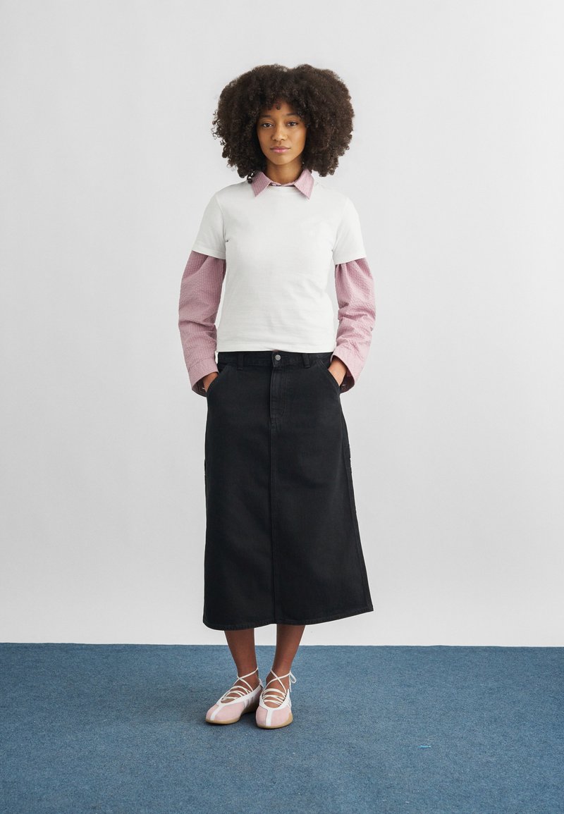 Donna con capelli ricci che indossa maglie bianche e rosa a maniche lunghe sovrapposte, gonna midi nera e ballerine rosa, in piedi su un tappeto blu.