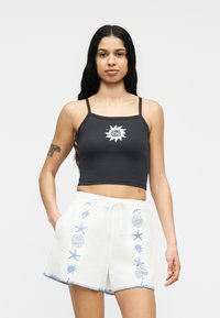 Γυναίκα που φοράει μαύρο κοντό τοπ με το λογότυπο του ήλιου «Santa Cruz» και λευκά σορτς με μπλε σχέδια θαλάσσιων πλασμάτων, στέκεται με τα χέρια στις τσέπες.