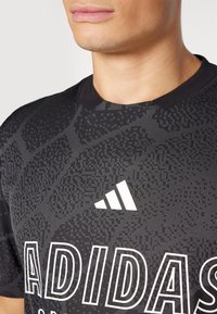 Camisola atlética preta com padrões texturizados, decote redondo e um logótipo branco da Adidas com letras em negrito na frente.