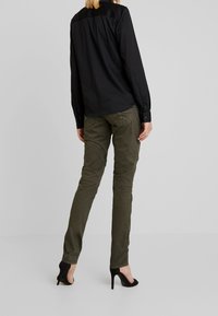 Maglietta nera a maniche lunghe con una texture liscia, abbinata a pantaloni verdi oliva slim-fit con dettagli di cucitura e indossata con tacchi alti neri.