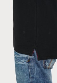 Chemise noire à manches courtes avec une fente latérale, associée à un jean bleu clair. La chemise présente un tissu texturé et un accent rouge subtil au niveau de la fente.
