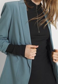 Ljusblå kavaj med en slät yta, öppen framsida och strukturerade axlar, lagd över en svart ribbad polotröja med dragkedja.