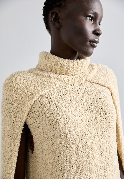 Beige textured sweater with a high collar, featuring a loose design and side openings. Material appears soft and fluffy, accentuating warmth.