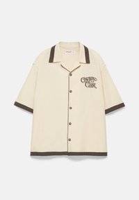 Chemise beige à manches courtes avec col, poignets et ourlet marron foncé, patte de boutonnage devant, et texte "Cowboy Code" sur la poitrine gauche.